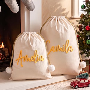 May include: Two off-white drawstring gift bags made of fabric, each featuring a yellow cursive name embroidered on the front. The smaller bag displays the name Amelia, while the larger bag displays the name Camila. Both bags have gold rope drawstrings with white pom-pom ends.