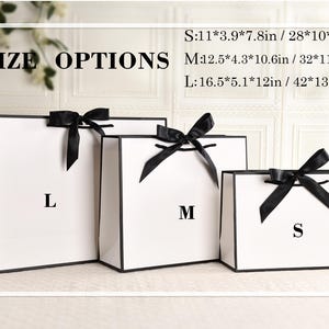 May include: Three white gift bags with black trim and black ribbon bows. The bags are labeled S, M, and L, with size options listed in inches and centimeters. The text "SIZE OPTIONS" is at the top.