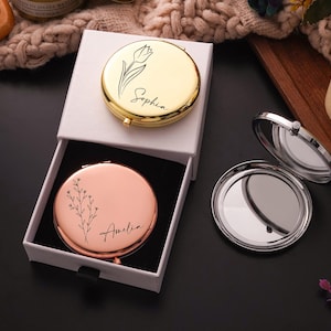 May include: Two personalized compact mirrors, one gold and one rose gold, are displayed in a white box. The gold mirror features a tulip design and the name "Sophia." The rose gold mirror has a floral design and the name "Amelia."