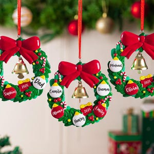 K&ouml;nnte beinhalten: Drei kreisf&ouml;rmige Weihnachtsbaumschmuck-Kr&auml;nze, die an roten B&auml;ndern h&auml;ngen. Jeder gr&uuml;ne Kranz hat eine gro&szlig;e rote Schleife oben, eine goldene Glocke in der Mitte und kleine rote und wei&szlig;e Kugeln, die mit Namen wie Mom, Dad und Camila beschriftet sind.