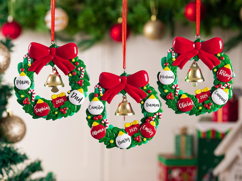 K&ouml;nnte beinhalten: Drei kreisf&ouml;rmige Weihnachtsbaumschmuck-Kr&auml;nze, die an roten B&auml;ndern h&auml;ngen. Jeder gr&uuml;ne Kranz hat eine gro&szlig;e rote Schleife oben, eine goldene Glocke in der Mitte und kleine rote und wei&szlig;e Kugeln, die mit Namen wie Mom, Dad und Camila beschriftet sind.