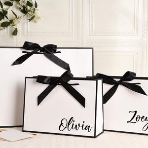 May include: Three white gift bags with black trim and black satin bows. The bags are different sizes, with the names "Olivia" and "Zoey" printed on two of them. The bags are on a white surface.