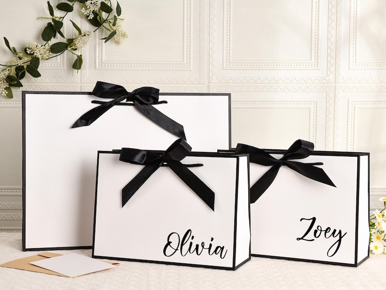 May include: Three white gift bags with black trim and black satin bows. The bags are different sizes, with the names "Olivia" and "Zoey" printed on two of them. The bags are on a white surface.
