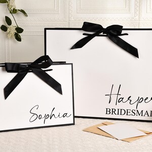 May include: Two white gift bags with black trim and black satin bows. One bag has the name "Sophia" in script, the other says "Harper Bridesmaid". A small card and envelope are in front.