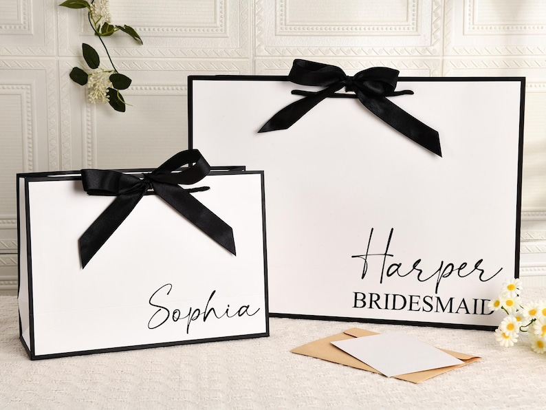 May include: Two white gift bags with black trim and black satin bows. One bag has the name "Sophia" in script, the other says "Harper Bridesmaid". A small card and envelope are in front.