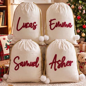 May include: Four off-white fabric drawstring bags featuring gold rope cords with white pom-poms at the ends. Each bag displays a name in red cursive script: Lucas, Emma, Samuel, and Asher, applied to the front of the canvas-like material.