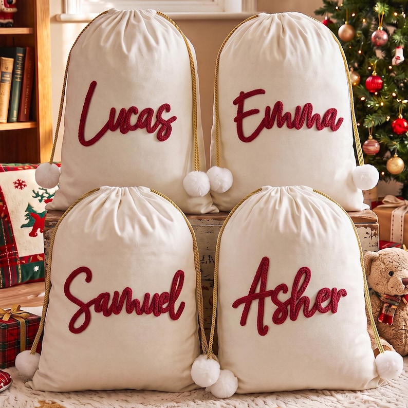 May include: Four off-white fabric drawstring bags featuring gold rope cords with white pom-poms at the ends. Each bag displays a name in red cursive script: Lucas, Emma, Samuel, and Asher, applied to the front of the canvas-like material.