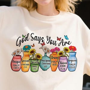 May include: Cream-colored sweatshirt with the text "God Says You Are" and colorful vases filled with flowers. Each vase has a word like "Unique" and a Bible verse reference. Butterflies are also featured.