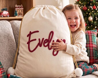 May include: A large, off-white fabric drawstring gift sack featuring the name Evelyn embroidered in thick, dark red yarn lettering. The sack has gold-colored braided drawstrings with white pom-poms at the ends, designed for holding holiday gifts.