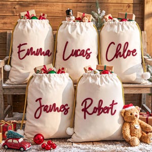 May include: Five cream-colored fabric drawstring sacks with gold cords and white pom-poms, each featuring a name embroidered in red script: Emma, Lucas, Chloe, James, and Robert. The sacks are filled with wrapped gifts and holiday ornaments.