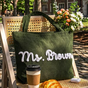 May include: A dark olive green corduroy tote bag featuring the white embroidered text Ms. Brown on the front. The bag has two matching fabric handles and is filled with a bouquet of white daisies and peach roses, accompanied by a coffee cup and croissant.