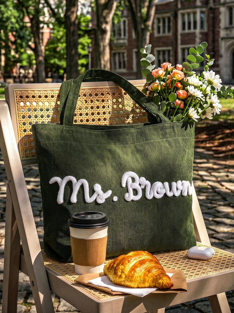 May include: A dark olive green corduroy tote bag featuring the white embroidered text Ms. Brown on the front. The bag has two matching fabric handles and is filled with a bouquet of white daisies and peach roses, accompanied by a coffee cup and croissant.