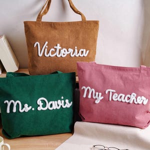 May include: Three corduroy tote bags in brown, green, and pink. Each bag features white, raised, cursive lettering. The brown bag reads Victoria, the green bag reads Ms. Davis, and the pink bag reads My Teacher. Each bag has two top handles.