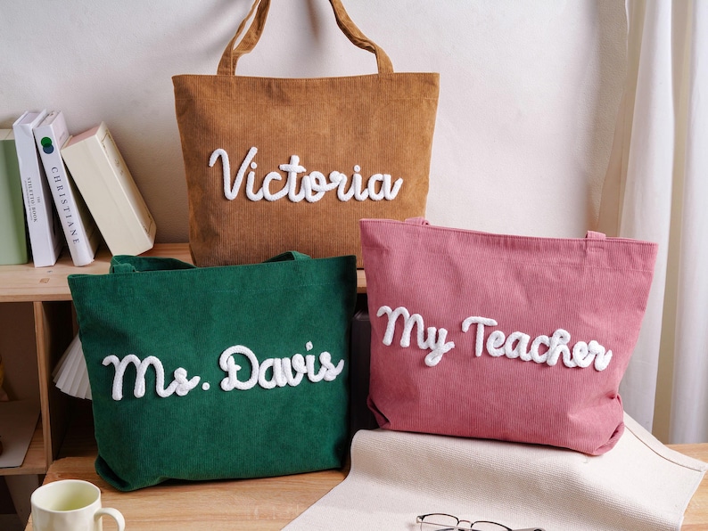 May include: Three corduroy tote bags in brown, green, and pink. Each bag features white, raised, cursive lettering. The brown bag reads Victoria, the green bag reads Ms. Davis, and the pink bag reads My Teacher. Each bag has two top handles.