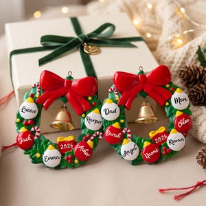 K&ouml;nnte beinhalten: Zwei kreisf&ouml;rmige Weihnachtsbaumschmuck-Kr&auml;nze mit gr&uuml;nem Blattwerk, einer gro&szlig;en roten Schleife oben und einer goldenen Glocke in der Mitte. Jeder Kranz ist mit kleinen roten und wei&szlig;en Kugeln verziert, die mit Namen wie Lucas, Jack, Emma, Harper, Asher, Dad, Sarah, Angel, Naomi, Mom, Chloe und Rowan sowie einem roten Geschenk mit der Aufschrift 2026 versehen sind.