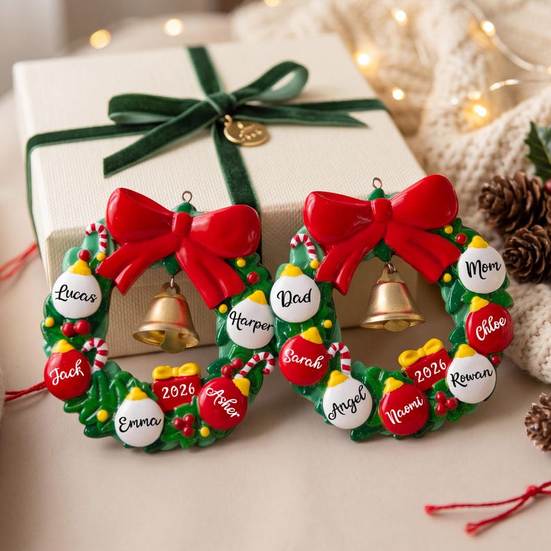 K&ouml;nnte beinhalten: Zwei kreisf&ouml;rmige Weihnachtsbaumschmuck-Kr&auml;nze mit gr&uuml;nem Blattwerk, einer gro&szlig;en roten Schleife oben und einer goldenen Glocke in der Mitte. Jeder Kranz ist mit kleinen roten und wei&szlig;en Kugeln verziert, die mit Namen wie Lucas, Jack, Emma, Harper, Asher, Dad, Sarah, Angel, Naomi, Mom, Chloe und Rowan sowie einem roten Geschenk mit der Aufschrift 2026 versehen sind.