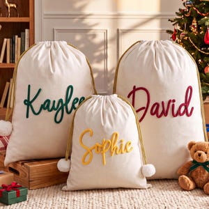 May include: Three off-white drawstring gift bags with gold rope cords and white pom-poms. The bags feature embroidered names in cursive script: Kaylee in dark green, David in red, and Sophie in yellow. The bags are made of fabric.