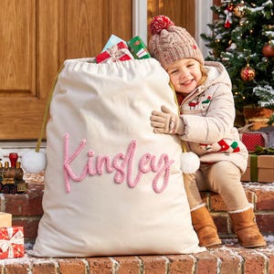 May include: A large, off-white fabric gift sack featuring the name Kinsley embroidered in pink yarn on the front. The sack has gold drawstring cords with white pom-poms at the ends and is filled with wrapped presents at the top opening.