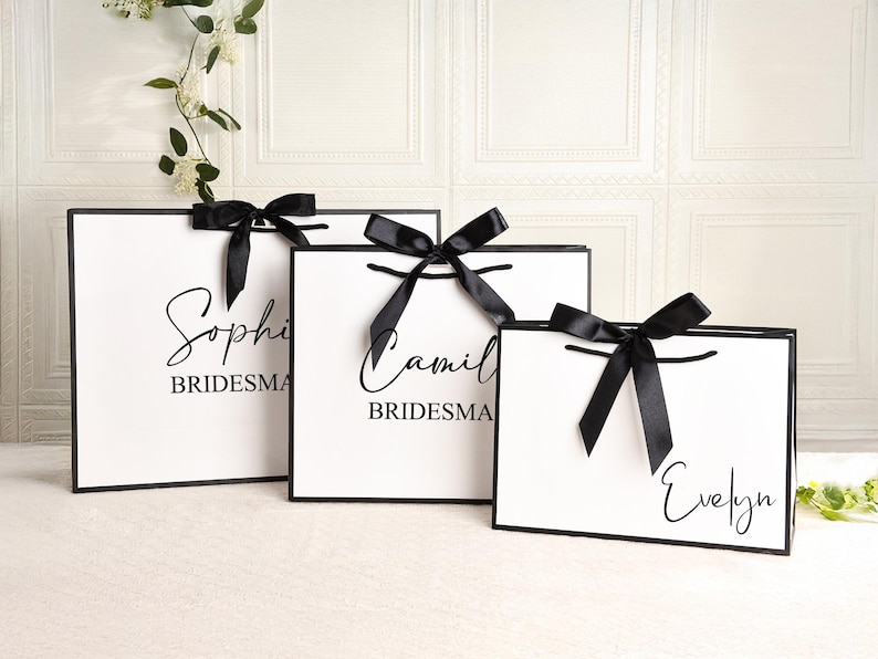 May include: Three white gift bags with black trim and black satin bows. Each bag has a name printed in black script: Sophie, Camila, and Evelyn. The word "Bridesmaid" is printed below the names.