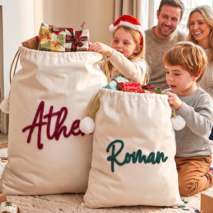 May include: Two off-white canvas drawstring gift sacks with gold cord ties and white pom-poms. The larger sack features the name Athena in dark red script, and the smaller sack features the name Roman in dark green script.