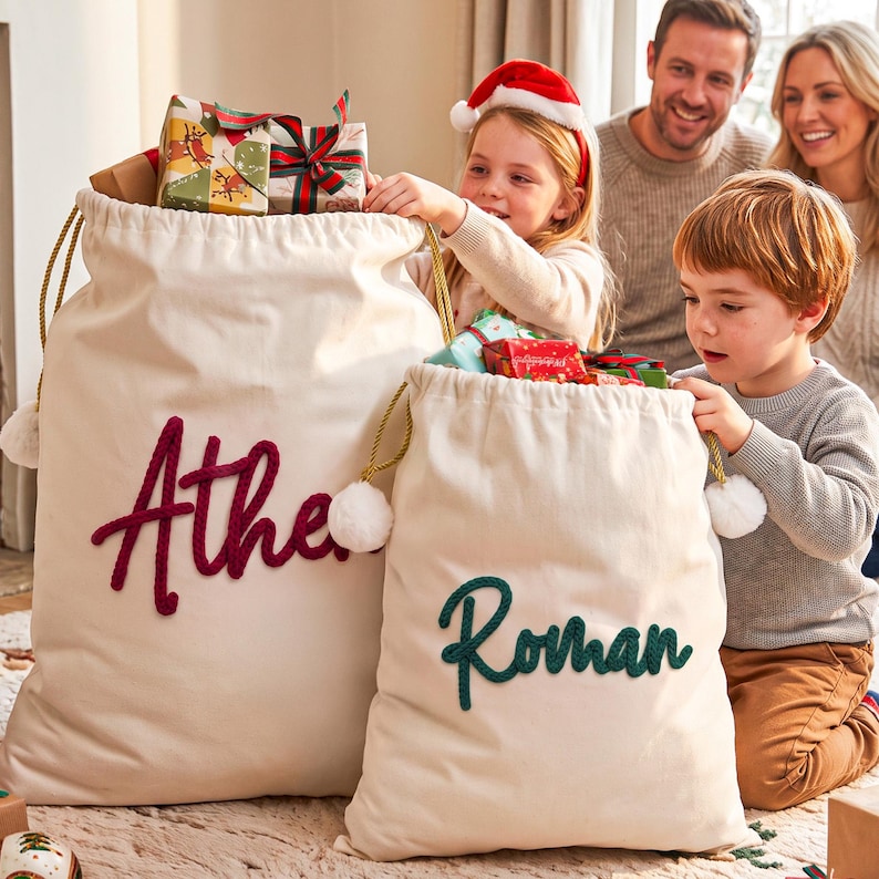 May include: Two off-white canvas drawstring gift sacks with gold cord ties and white pom-poms. The larger sack features the name Athena in dark red script, and the smaller sack features the name Roman in dark green script.