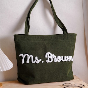 May include: A dark olive green corduroy tote bag featuring two shoulder straps and the text "Ms. Brown" embroidered in white, raised, cursive lettering across the front center of the bag, showcasing a textured fabric surface.
