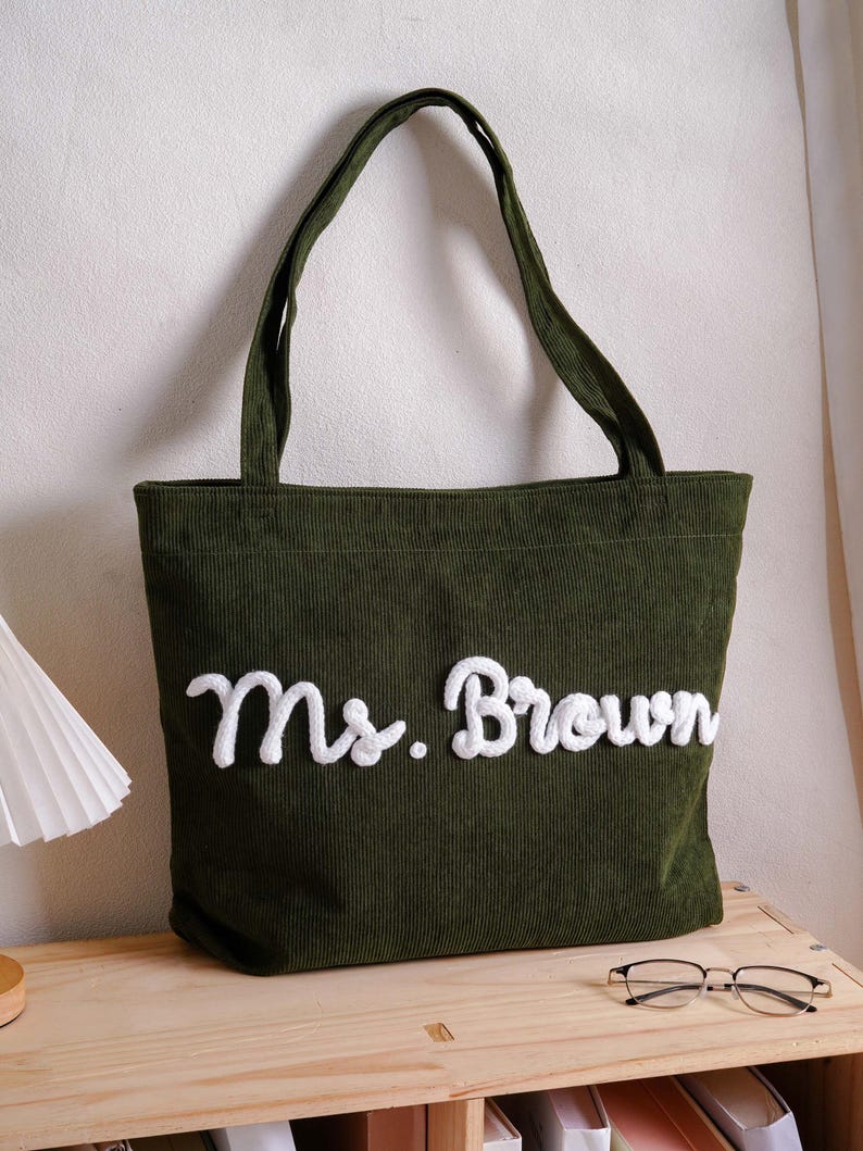May include: A dark olive green corduroy tote bag featuring two shoulder straps and the text "Ms. Brown" embroidered in white, raised, cursive lettering across the front center of the bag, showcasing a textured fabric surface.