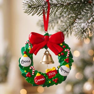 K&ouml;nnte beinhalten: Ein gr&uuml;ner Weihnachtskranz-Anh&auml;nger mit einer gro&szlig;en roten Schleife oben und einer goldenen Glocke in der Mitte. Der Kranz ist mit roten Beeren, Zuckerstangen und drei wei&szlig;en oder roten Kugeln verziert, die mit den Namen Lucas, Emma und Harper sowie einem kleinen roten Geschenk mit der Aufschrift 2026 versehen sind.