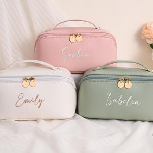 Puede incluir: Tres neceseres personalizados en colores pastel: rosa, blanco y verde salvia. Cada neceser tiene un asa, una cremallera dorada y un nombre en escritura cursiva. Los neceseres están colocados sobre una superficie blanca con flores al fondo.