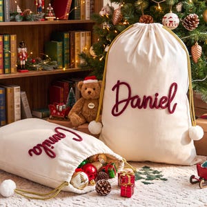 May include: Two off-white canvas drawstring bags featuring gold braided cords with white pom-poms at the ends. One bag is upright and displays the name Daniel in raised red script, while the other lies open, revealing the name Camila in red.