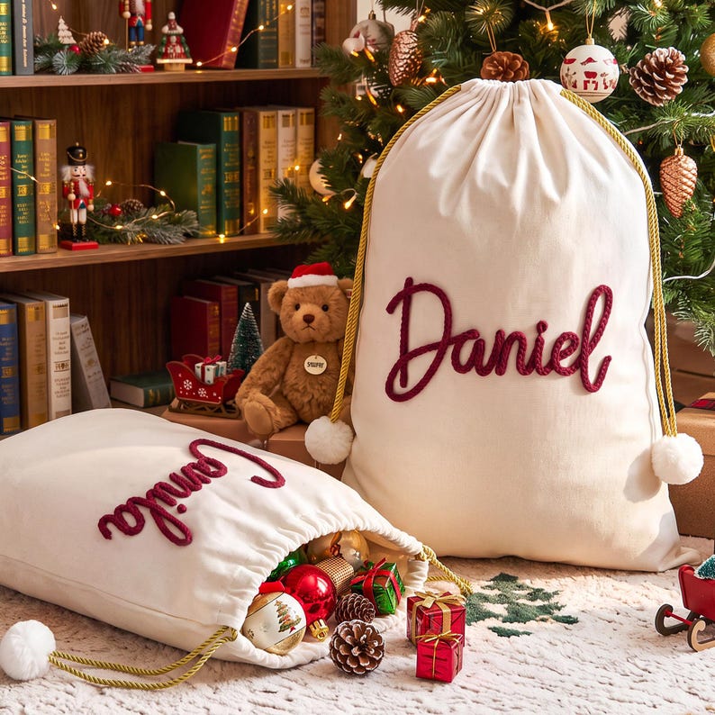 May include: Two off-white canvas drawstring bags featuring gold braided cords with white pom-poms at the ends. One bag is upright and displays the name Daniel in raised red script, while the other lies open, revealing the name Camila in red.