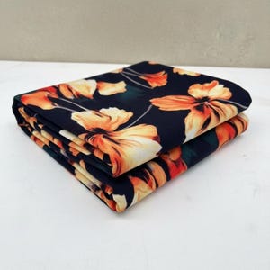 May include: Folded fabric with a floral pattern. The fabric is dark blue with large orange and yellow flowers and green leaves. The fabric is folded neatly in a stack, showing the floral design.