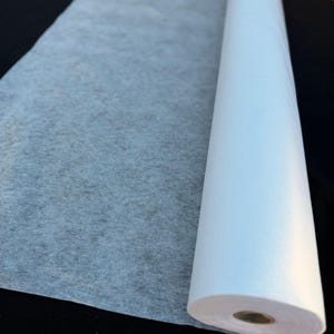 Puede incluir: Un rollo de tela de entretela blanca, semitranslúcida y no tejida. El material es fino y fibroso, con una textura visible que permite el paso de la luz. El rollo es cilíndrico y cuenta con un núcleo de cartón en el centro.
