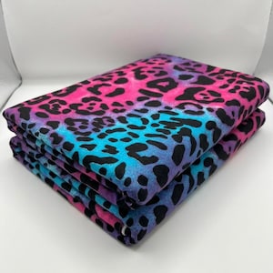 Peut inclure: Une pile de tissu plié présentant un motif léopard vibrant. Le textile affiche un fond dégradé aux teintes rose vif, violet et bleu, recouvert d'un motif répétitif de taches d'animaux noires sur toute la surface du tissu.