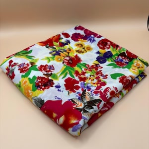 May include: A folded square of white fabric featuring a vibrant, multi-coloured floral pattern. The design includes large red, yellow, and purple blossoms interspersed with green leaves and smaller floral accents across the entire surface.