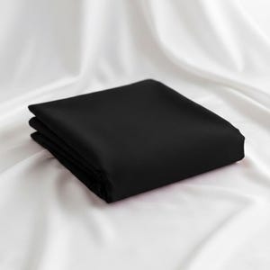 Può includere: Un tessuto nero quadrato e piegato, forse un lenzuolo o un panno, è presentato su uno sfondo di tessuto bianco. Il tessuto nero è ben piegato, con una superficie liscia e opaca. Il contrasto tra il nero e il bianco crea una presentazione visivamente suggestiva.