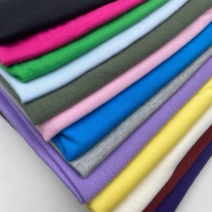May include: A stack of folded fabric pieces in a variety of solid colours including black, hot pink, green, light blue, olive, light pink, bright blue, grey, lavender, yellow, cream, maroon, and dark purple, all arranged in a neat, layered pile.