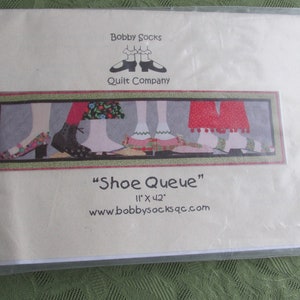 Op de afbeelding: Een witte kaart met de tekst "Bobby Socks Quilt Company" en een afbeelding van een rij kleurrijke schoenen met de tekst "Shoe Queue" en "11" x "42" eronder. Het websiteadres "www.bobbysocksqc.com" staat onderaan de kaart gedrukt.