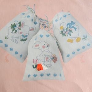 May include: Three white fabric drawstring bags with embroidered designs of a bunny, a goose, and a bunny holding a basket. The bags have blue and white trim.