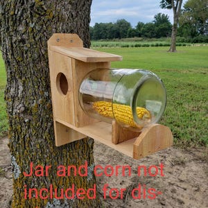 May include: A wooden bird feeder with a clear glass jar attached, mounted on a tree. The feeder has a small perch and a circular opening. The jar is designed to hold birdseed or corn. The text on the image says "Jar and corn not included for display purposes."