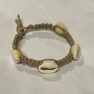 May include: A woven bracelet made from a tan-coloured cord, decorated with five white and cream-coloured seashells. The bracelet is knotted and adjustable. The shells are smooth and oval-shaped, creating a nautical theme.