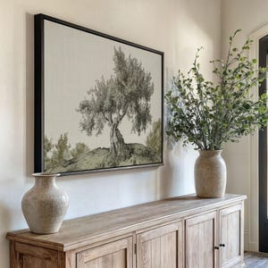 Peut inclure: Une œuvre d'art encadrée représentant un paysage d'arbres en monochrome. L'œuvre est suspendue au-dessus d'un buffet en bois, avec un vase sur le buffet et un autre vase avec de la verdure à droite. La pièce a une palette de couleurs neutres.