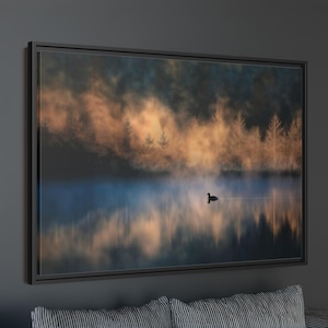 May include: A framed canvas print depicts a serene lake scene. The water reflects the sky and trees, with a lone duck swimming. The sky is a mix of blue and orange, with a misty effect. The artwork is framed in a black border.