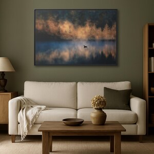 May include: A framed landscape art print depicts a serene lake scene. The artwork features a misty atmosphere with a duck on the water, and the reflection of trees. The color palette includes blues, oranges, and greens, creating a tranquil ambiance.