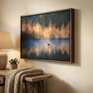 May include: A framed canvas art print depicts a serene lake scene with a single bird. The artwork features a misty atmosphere with golden and blue hues, and is encased in a dark wooden frame. The artwork is mounted on a beige wall.