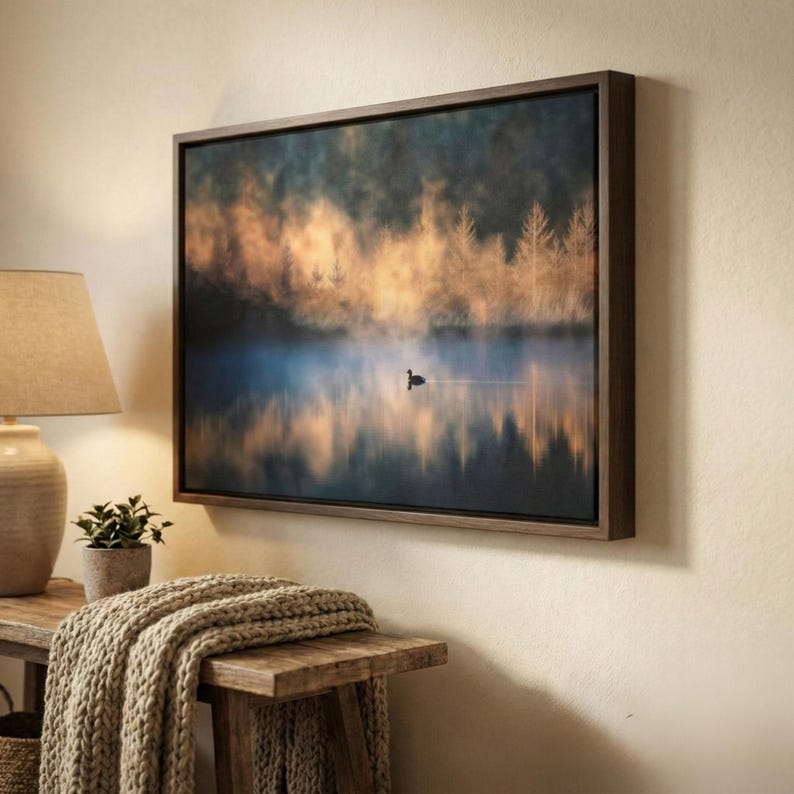 May include: A framed canvas art print depicts a serene lake scene with a single bird. The artwork features a misty atmosphere with golden and blue hues, and is encased in a dark wooden frame. The artwork is mounted on a beige wall.