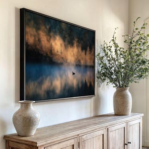 May include: A framed landscape art print depicts a serene lake scene with a lone bird. The artwork features a misty atmosphere with golden and blue hues, reflecting in the water. The print is displayed above a wooden cabinet, with two decorative vases and greenery.