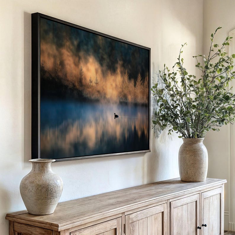 May include: A framed landscape art print depicts a serene lake scene with a lone bird. The artwork features a misty atmosphere with golden and blue hues, reflecting in the water. The print is displayed above a wooden cabinet, with two decorative vases and greenery.
