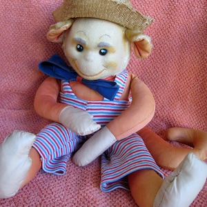 May include: A plush monkey toy wearing a straw hat, blue bow tie, and red and white striped overalls. The monkey is sitting on a pink fuzzy surface.