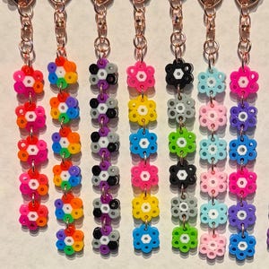 May include: A selection of keyrings with flower-shaped charms crafted from colourful beads. Each keyring displays a unique colour palette, including pink, blue, green, and yellow. The charms are connected to copper-coloured metal chains.